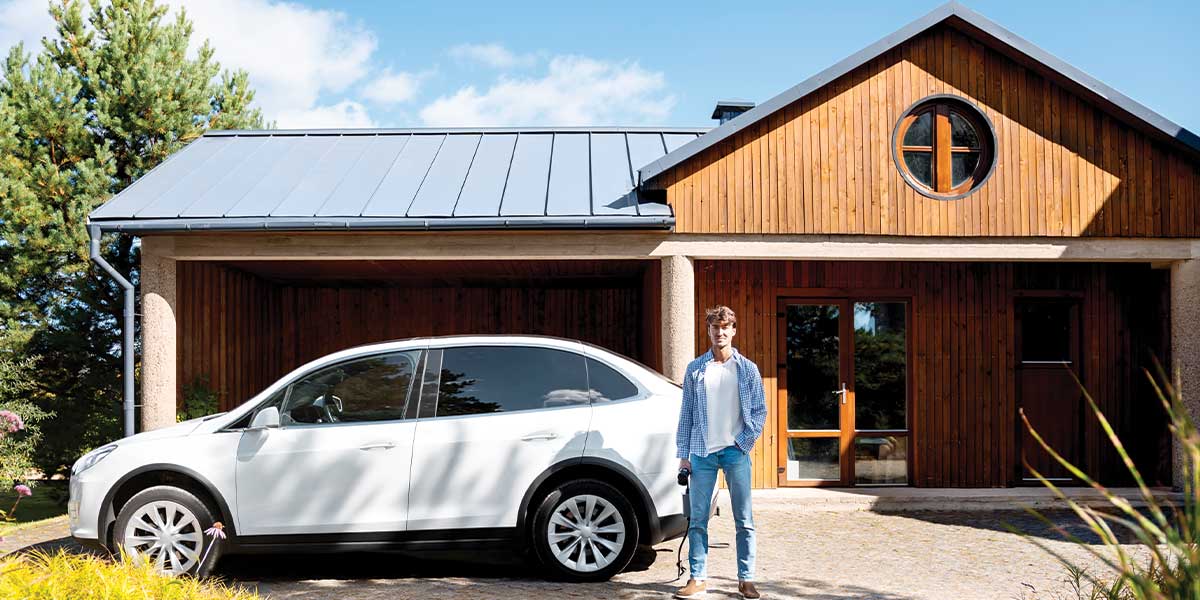 Person standing in front of their electric car with their home in the background on a sunny day.