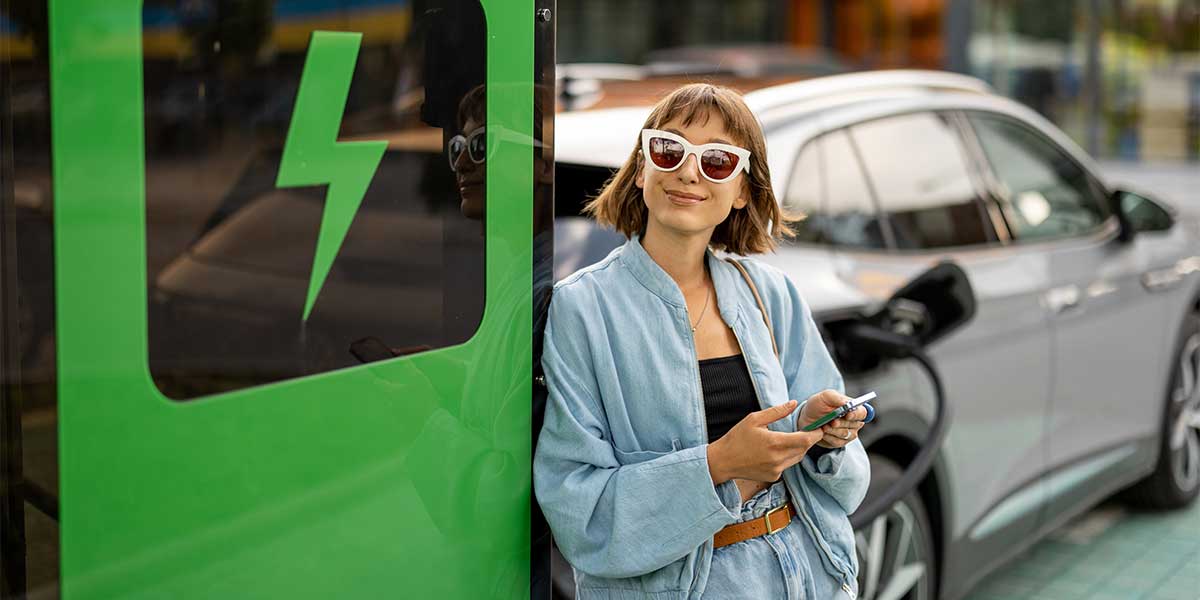Woman relaxing next to her electric vehicle at a charging station, representing the lifestyle shift addressed in Electric Vehicle Myths Debunked—where EV charging is convenient, quick, and part of daily routine.