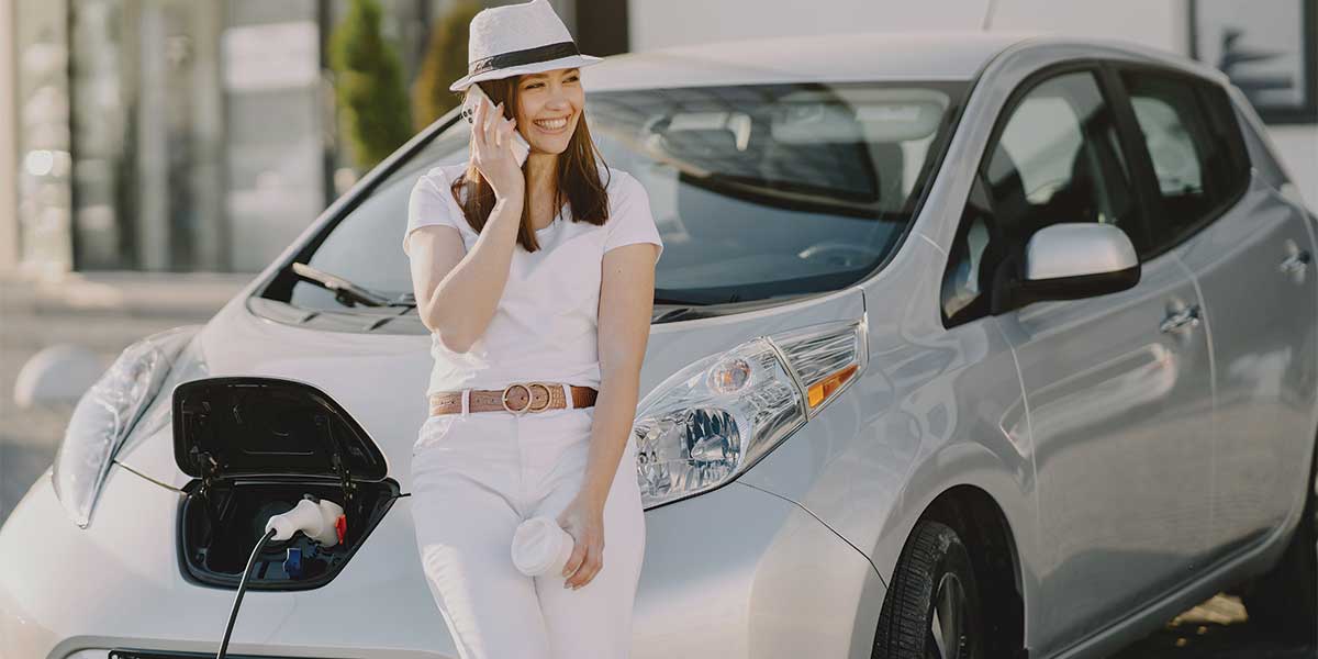 A woman standing next to a charging electric car, highlighting the growing accessibility and usability of EVs in the Evolution of Electric Vehicles.