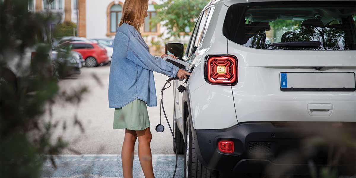 A woman is standing next to a white electric vehicle (EV), plugging it into a charging station. - Top 5 EV Charger Brands