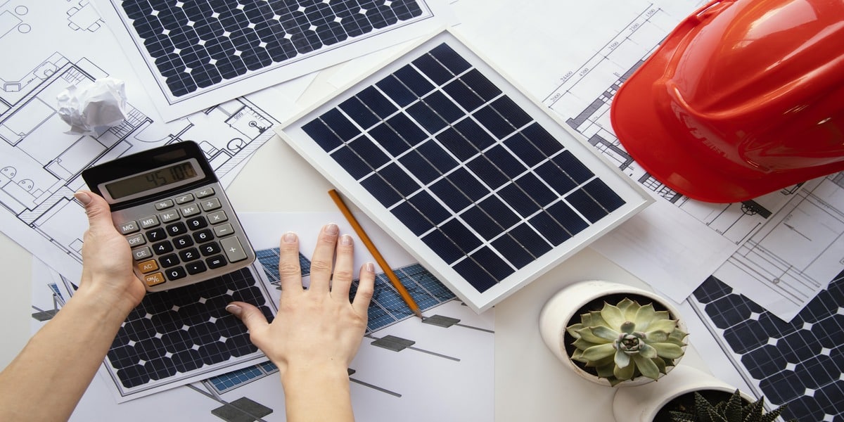 Hands holding a calculator over solar panel models and blueprints, surrounded by tools and a red hard hat, showcasing the planning stage of a solar installation. Ideal for conveying Expert Electric’s role in applying 2025 solar incentives and rebates to reduce project costs.
