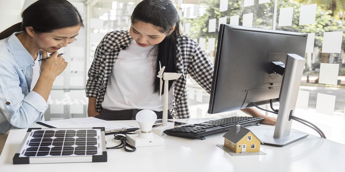 Two women at an office desk reviewing documents beside a solar panel model and a desktop monitor, exploring residential solar energy options. This image visually supports Expert Electric’s mission to help homeowners understand solar incentives and rebates in 2025.