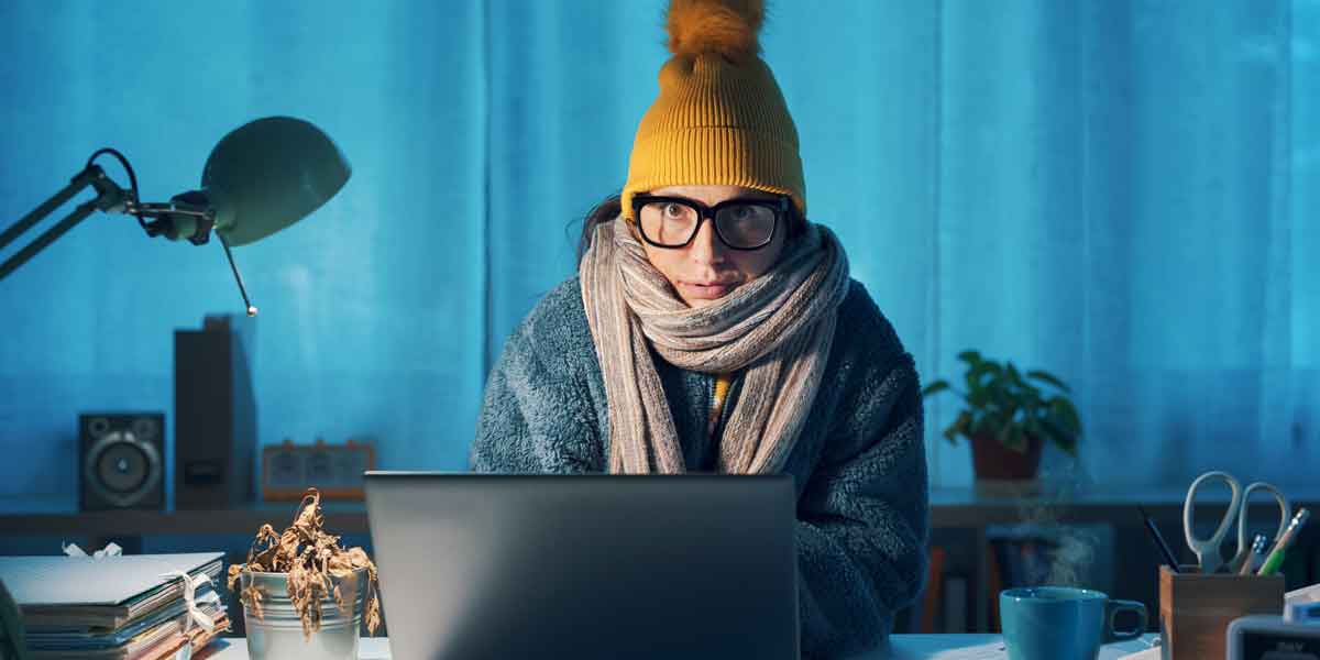 Woman in a yellow toque and a winter jacket sits in her home cold while working on a laptop.