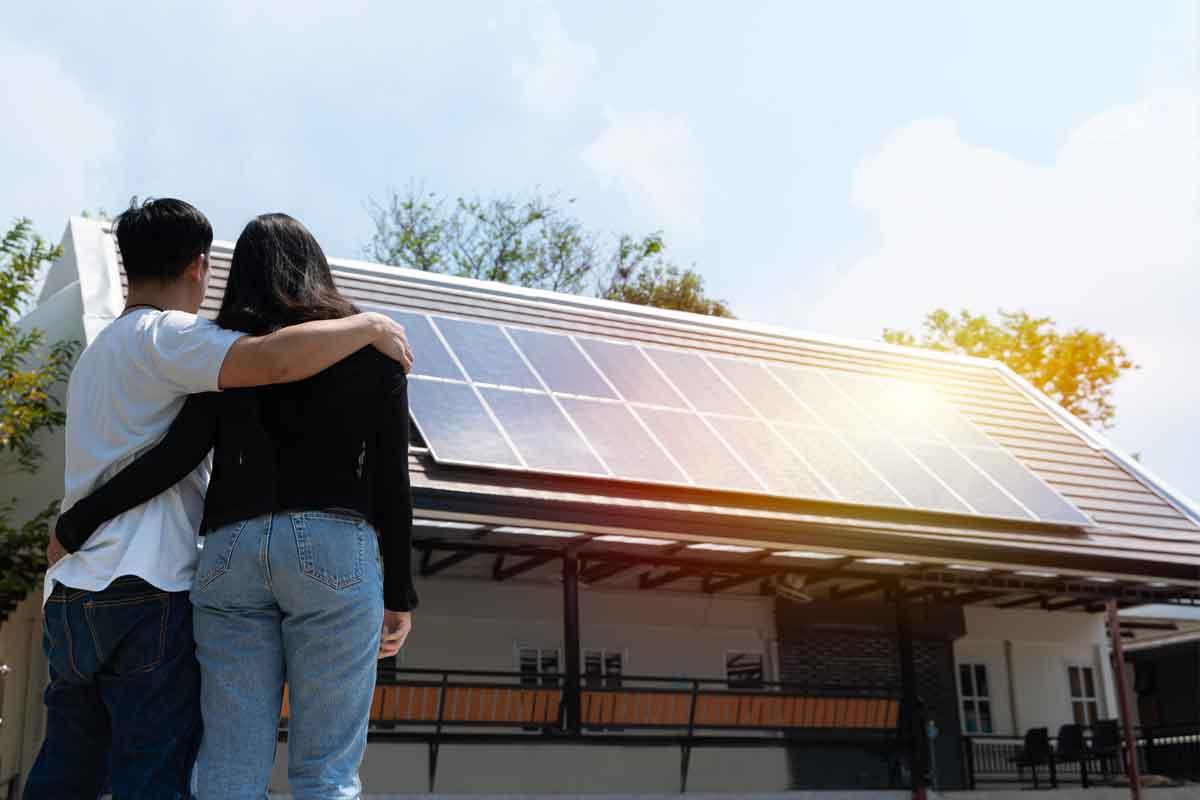 A couple is standing in the foreground, proud, while looking at the solar panels installed on their roof on a sunny day.